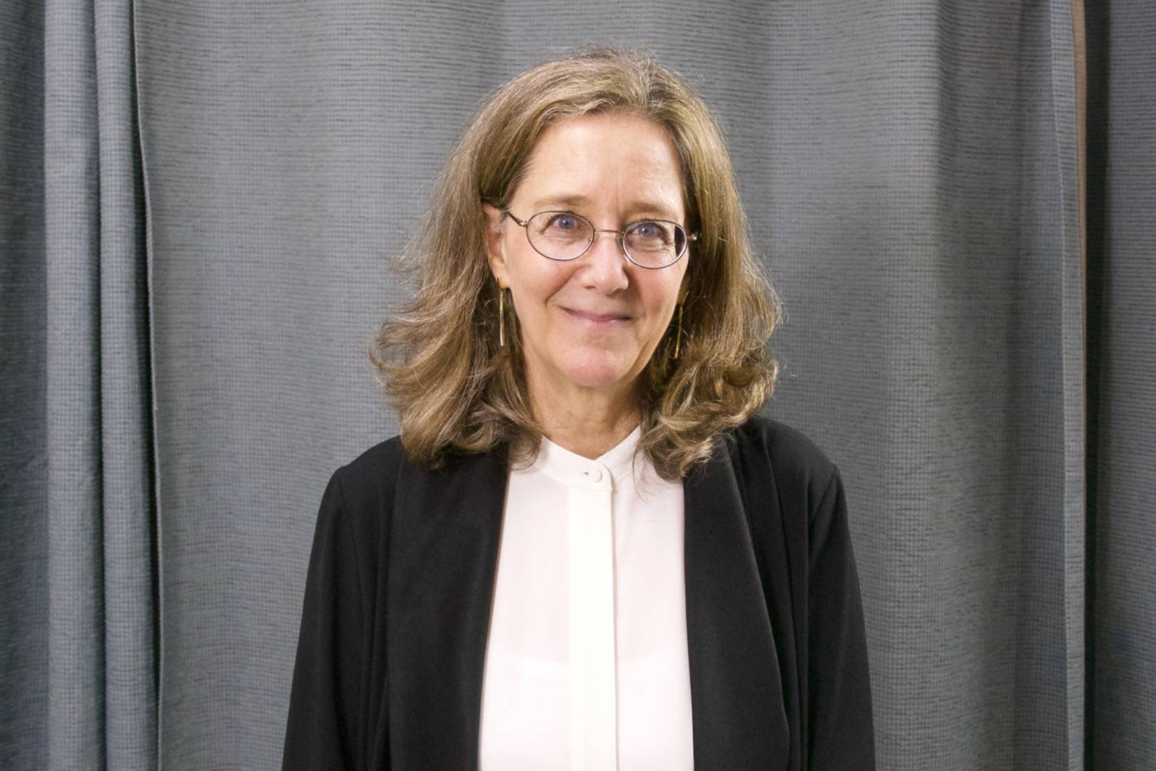 A woman in glasses and black jacket standing next to a curtain.