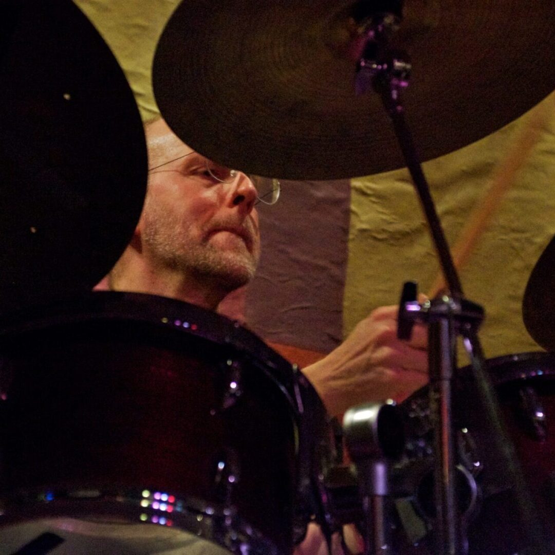 A man playing drums in front of a yellow wall.