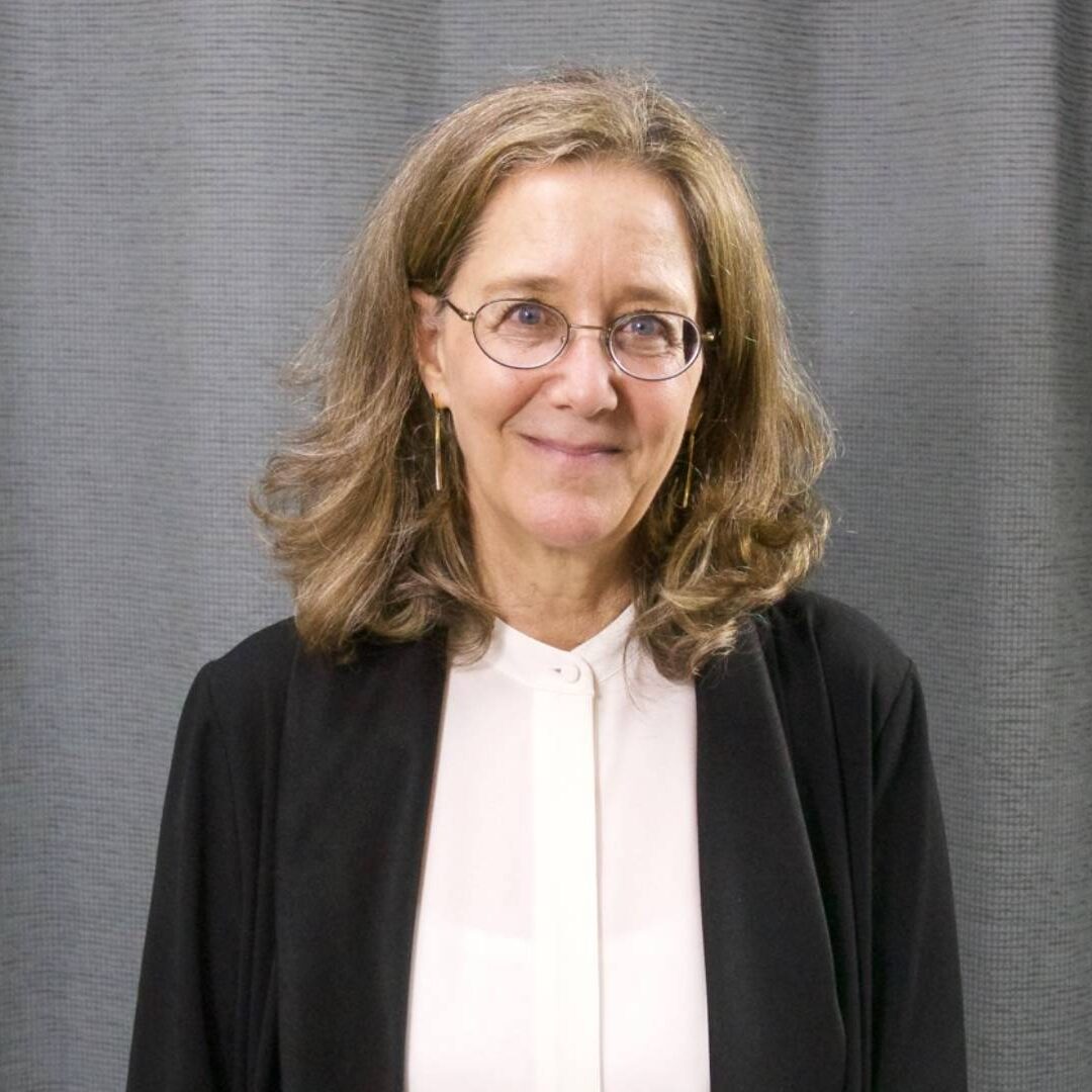 A woman in glasses and black jacket standing next to a curtain.